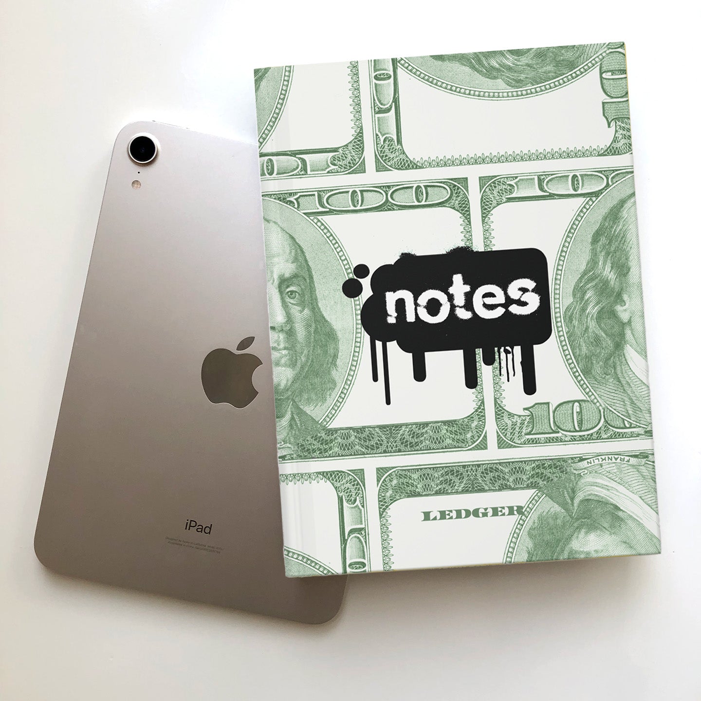 Apple iPad next to a notebook with a 100 dollar bill design and 'notes' sticker-like graphic.