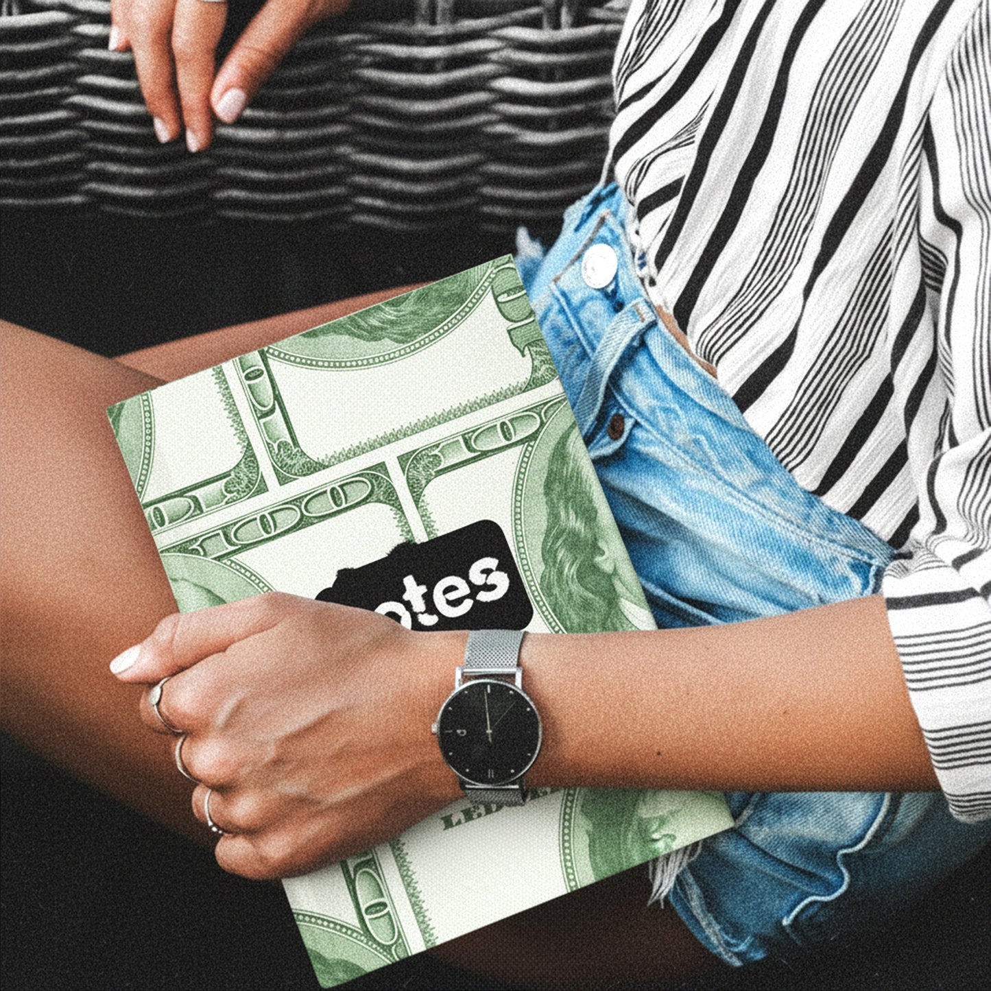 Woman holding a notebook, a watch on their wrist, wearing a striped shirt and denim shorts.