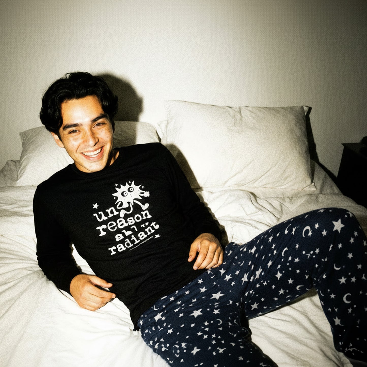 Person wearing a black shirt and star-patterned pants sitting on a bed.
